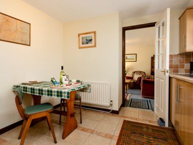 A small dining area with a wooden table covered by a green checkered tablecloth and two chairs next to a kitchen with wooden cabinets and a tiled floor at Swallows Cottage nr Warkworth in Acklington