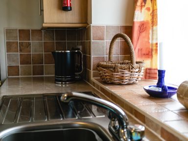 A kitchen sink with a faucet and a black electric kettle next to a woven basket and a blue candle holder on the tiled countertop at Swallows Cottage nr Warkworth in Acklington
