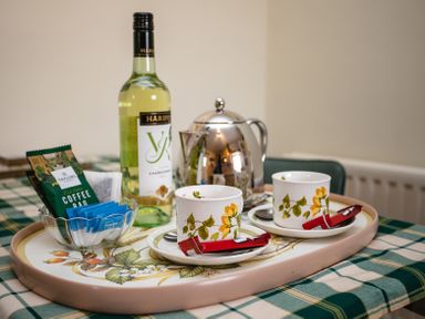 A tray with two cups and saucers, a metal teapot, a bottle of white wine, coffee bags, and sugar packets on a checkered tablecloth at Swallows Cottage nr Warkworth in Acklington