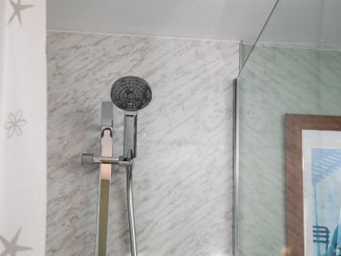 A showerhead attached to a metal fixture on a marble tiled wall with a glass panel and framed artwork in bathroom at Swallows Cottage nr Warkworth in Acklington