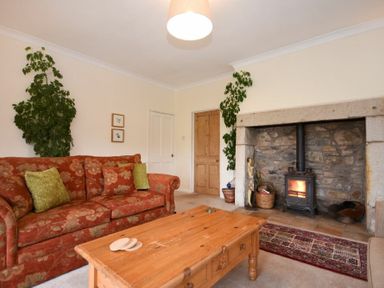 A living room with a floral sofa a wooden coffee table plants and a stone fireplace with a wood stove at Dunrovin in Beadnell