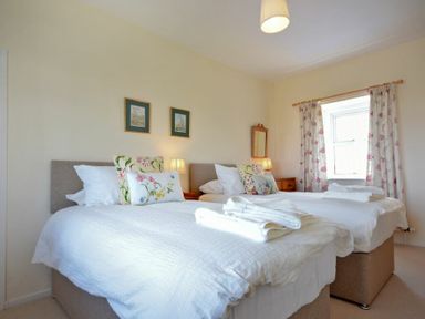A bedroom with two single beds white bedding floral cushions bedside tables lamps a window with floral curtains and two pictures on the wall at Dunrovin in Beadnell