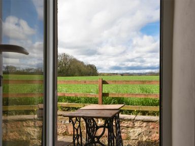 View through a glass door of a wooden table with a metal base outside with a wooden fence and green field in the background at Dunrovin Beadnell