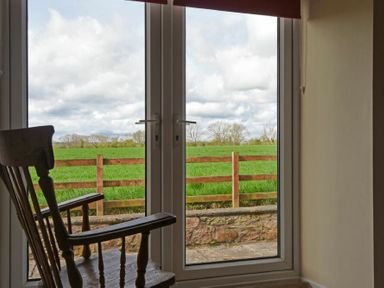 A wooden rocking chair next to glass doors with a view of a green field and wooden fence at Dunrovin in Beadnell