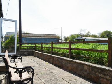 A stone patio with a wooden bench and metal arms next to a wooden fence and grassy field with farm buildings in the background at Dunrovin in Beadnell