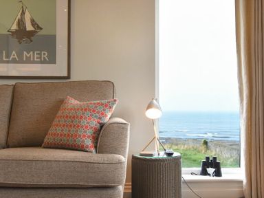 A sofa with a patterned cushion next to a small table with a lamp and a window showing a sea view at Portside in Seahouses