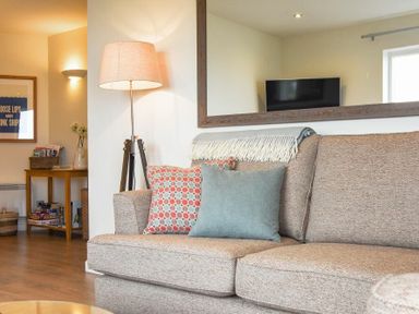 A living room with a beige sofa with cushions and a throw and a floor lamp next to a wooden table with books and magazines at Portside in Seahouses