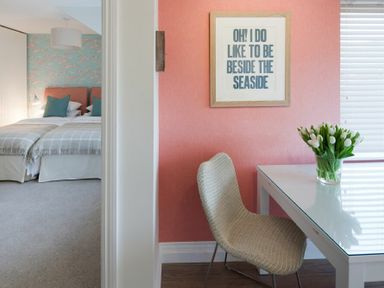 A bedroom with two single beds and a bed with pillows next to a room with a table, chair, flower vase, and framed text on a coral wall at Portside in Seahouses