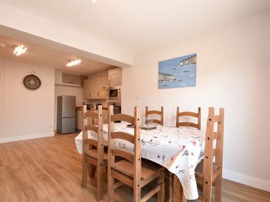 A dining area with a wooden table and six chairs with a tablecloth next to a kitchen with appliances and a wall clock at Anchor Lodge in Seahouses