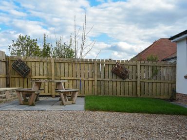 A backyard with a wooden picnic table and benches on stone tiles next to a grassy area and a wooden fence