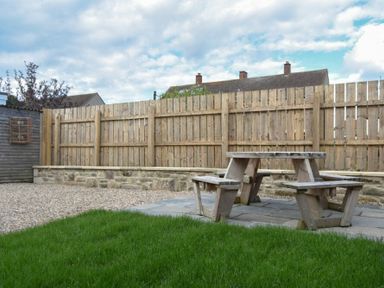 A wooden picnic table on a stone patio in a backyard with a grass lawn and wooden fence at Anchor Lodge in Seahouses