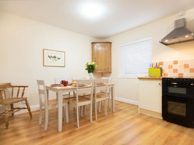 A kitchen with a dining table set with plates and cups a wooden chair a wall cabinet a window with blinds a stove and a range hood at Summerfold in Warkworth