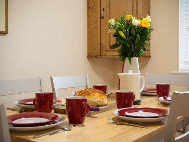 A dining table set with red and white plates and mugs with bread on a wooden board and a white pitcher with flowers in a vase at Summerfold in Warkworth