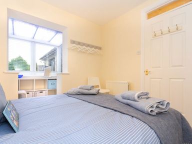 A bedroom with a blue bedspread, folded towels on the bed, a white chair, window with blinds and built-in storage at Summerfold in Warkworth