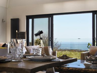 A dining area with a wooden table set with plates and glasses and a telescope by sliding glass doors overlooking the sea at Waters Edge Beadnell Harbour Road in Beadnell