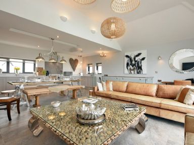 An open plan living and dining area with a brown sofa a pebble coffee table and a kitchen with a dining table and benches at Waters Edge Beadnell (Harbour Road) in Beadnell