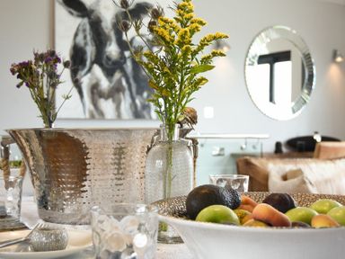 A table with a fruit bowl and flowers in a glass vase with a cow painting and round mirror in the background at Waters Edge Beadnell Harbour Road in Beadnell