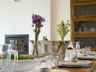 A dining table set with plates, glasses, flowers in vases, and a metal ice bucket in a room with a wooden cabinet and fireplace at Waters Edge Beadnell (Harbour Road) in Beadnell