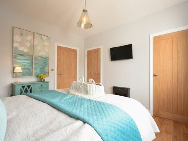 A bedroom with a bed covered with a white and blue quilt a blue cabinet with a lamp and flowers on top two wooden doors a wall-mounted tv and a hanging ceiling light at Waters Edge Beadnell Harbour Road in Beadnell