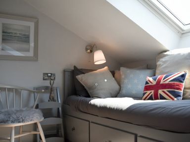 A small bedroom with a built-in bed under a skylight with cushions including a union jack pillow a wooden chair with a fur cushion and a wall lamp at Waters Edge Beadnell (Harbour Road) in Beadnell