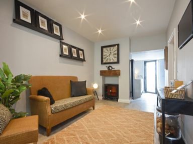 A sitting room with a brown sofa, a large wall clock, and framed pictures on shelves at Waters Edge Beadnell Harbour Road in Beadnell