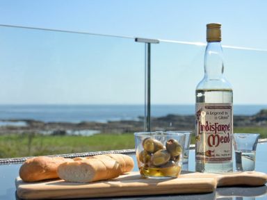 A bottle of Lindisfarne Mead with a glass of green olives and sliced bread on a wooden board outside with a coastal view at Waters Edge Beadnell Harbour Road in Beadnell