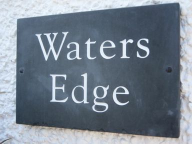 A black sign with white text reading Waters Edge mounted on a textured white wall at Waters Edge Beadnell (Harbour Road) in Beadnell