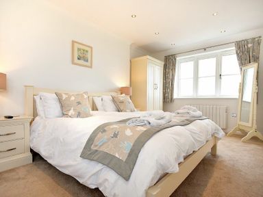 A bedroom with a double bed, bedside tables with lamps, a wardrobe, a standing mirror, and windows with curtains at Portland in Beadnell