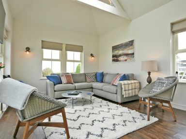A living room with a gray corner sofa, two woven chairs, a round coffee table, a white patterned rug, and wall art at South View nr Bamburgh in Seahouses
