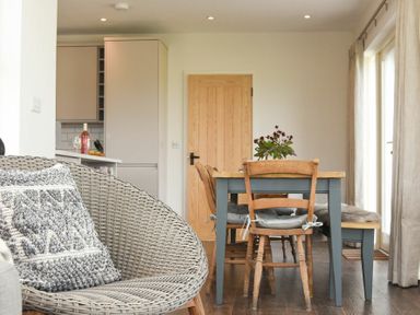 A dining area with wooden chairs and table a woven chair with cushion and kitchen in the background at South View nr Bamburgh in Seahouses