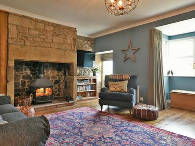 A living room with a stone fireplace a wood burning stove an armchair a patterned rug and a wooden chest by the window at South View nr Bamburgh in Seahouses