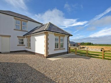 A white house with windows and a gravel driveway next to a wooden fence with fields beyond at South View nr Bamburgh in Seahouses