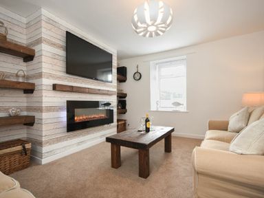 A living room with a wall-mounted TV above an electric fireplace a wooden coffee table a beige sofa and wooden shelves at Rivershield in Warkworth