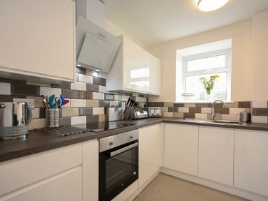 A kitchen with white cabinets a stove a sink a window with flowers on the sill and a tiled backsplash at Rivershield in Warkworth