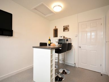 A small kitchen area with a breakfast bar, two black bar stools, a bottle of wine, a bread loaf, a wine glass, a microwave, and a wall-mounted TV at Rivershield in Warkworth