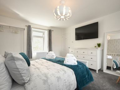 A bedroom with a bed folded white towels a dresser a wall mounted television a full length mirror and a window with curtains at Rivershield in Warkworth