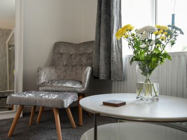 A grey armchair with matching footstool next to a white round table with a vase of yellow and white flowers at Rivershield in Warkworth