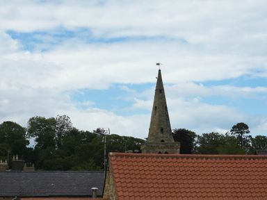 A stone church steeple with a weather vane behind rooftops with tile and slate roofs with trees and cloudy sky in the background at Rivershield in Warkworth