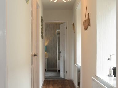 A hallway with wooden floor and white walls with a door at the end showing a room with wallpaper and a bedside table with a lamp at Rivershield in Warkworth