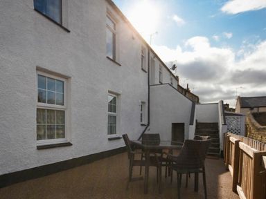 An outdoor patio area with a glass table and four chairs next to a white building with stairs leading up at Rivershield in Warkworth