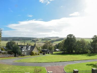 A rural landscape with a stone house trees grass and hills under a partly cloudy sky at The Lodge (Elsdon) in Elsdon