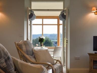 A seating area with armchairs and cushions next to a door leading to a sunroom with a table, flowers, and a view of hills at The Lodge (Elsdon) in Elsdon