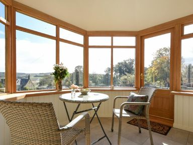 A sunroom with wicker chairs a round glass table and large windows showing outdoor trees at The Lodge Elsdon