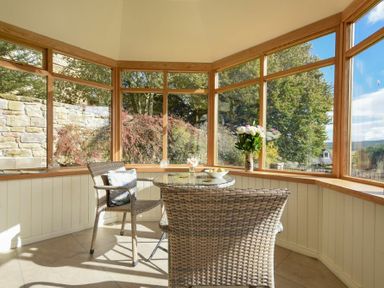 A sunroom with wicker chairs and a glass table surrounded by large windows at The Lodge (Elsdon) in Elsdon