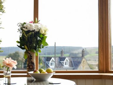 A vase with flowers and a bowl of fruit on a table by a window showing a house and hills outside at The Lodge Elsdon