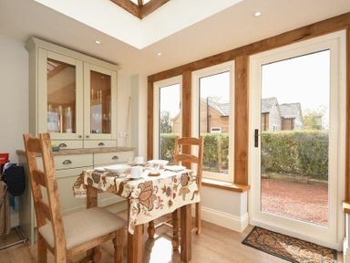 A dining area with a wooden table covered with a patterned tablecloth two wooden chairs a glass cabinet and large windows at The Lodge Elsdon