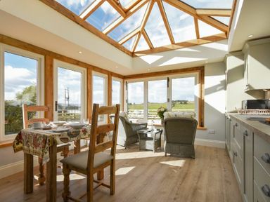A dining area with wooden table and chairs next to a seating area with wicker chairs under a glass ceiling at The Lodge (Elsdon) in Elsdon