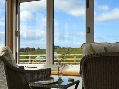 A seating area with two wicker chairs and a table in front of a large window showing a green field at The Lodge (Elsdon) in Elsdon