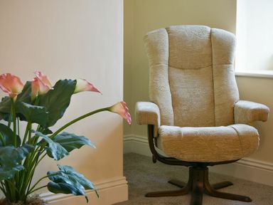A beige upholstered armchair with wooden base next to a potted plant with pink flowers at The Lodge (Elsdon) in Elsdon