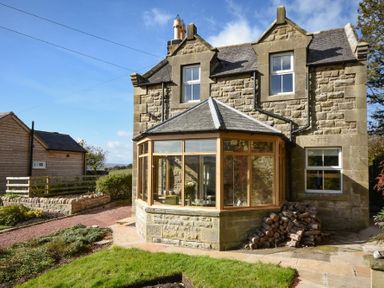 A stone house with a glass sunroom and stacked firewood outside at The Lodge (Elsdon) in Elsdon
